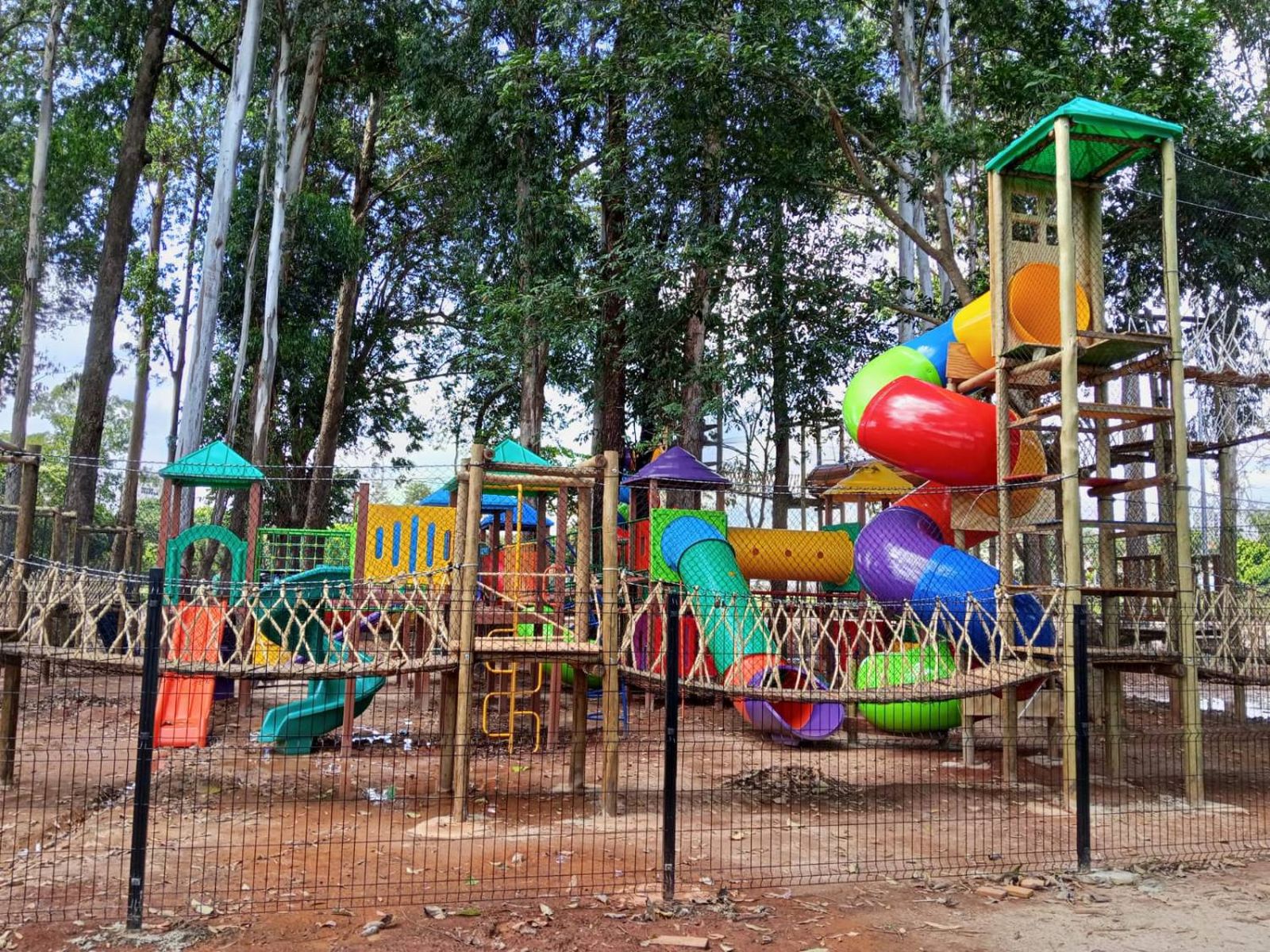 Kidsplay de madeira com torre, escorregadores e tubos coloridos, cercado por grade, entre árvores altas